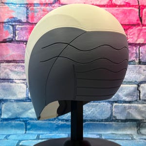 May include: A two-tone helmet, with a light beige upper section and a dark gray lower section. The dark gray portion features curved, wave-like lines. The helmet is displayed on a black stand against a colorful brick wall background.