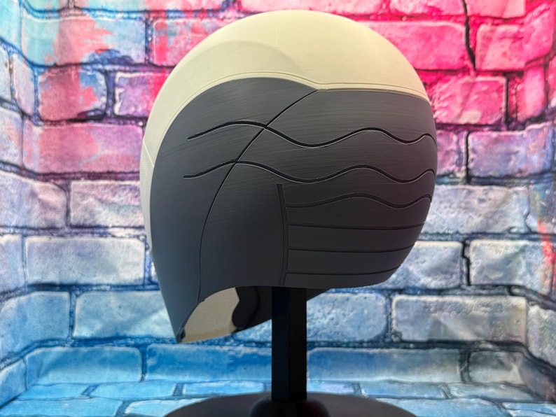 May include: A two-tone helmet, with a light beige upper section and a dark gray lower section. The dark gray portion features curved, wave-like lines. The helmet is displayed on a black stand against a colorful brick wall background.
