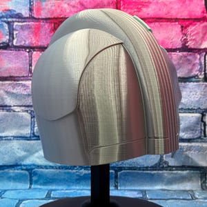 May include: A light gray helmet with a rounded top and a ridged back section. The helmet has a smooth, metallic appearance and is mounted on a black stand. The background features a brick wall with blue, pink, and white paint.