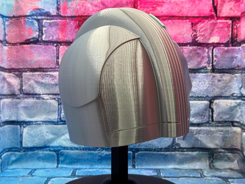 May include: A light gray helmet with a rounded top and a ridged back section. The helmet has a smooth, metallic appearance and is mounted on a black stand. The background features a brick wall with blue, pink, and white paint.