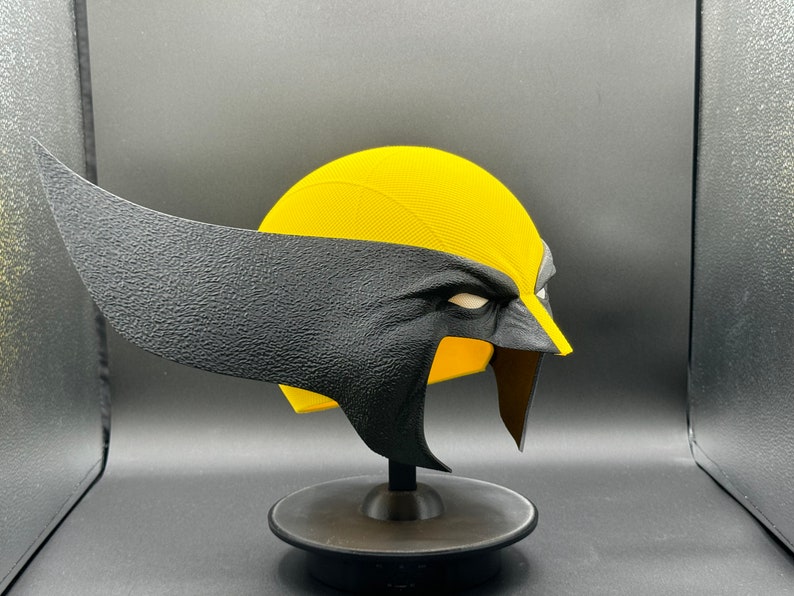 Puede incluir: Una m&aacute;scara de Wolverine de pl&aacute;stico amarillo y negro con una superficie texturizada. La m&aacute;scara est&aacute; en un soporte negro.