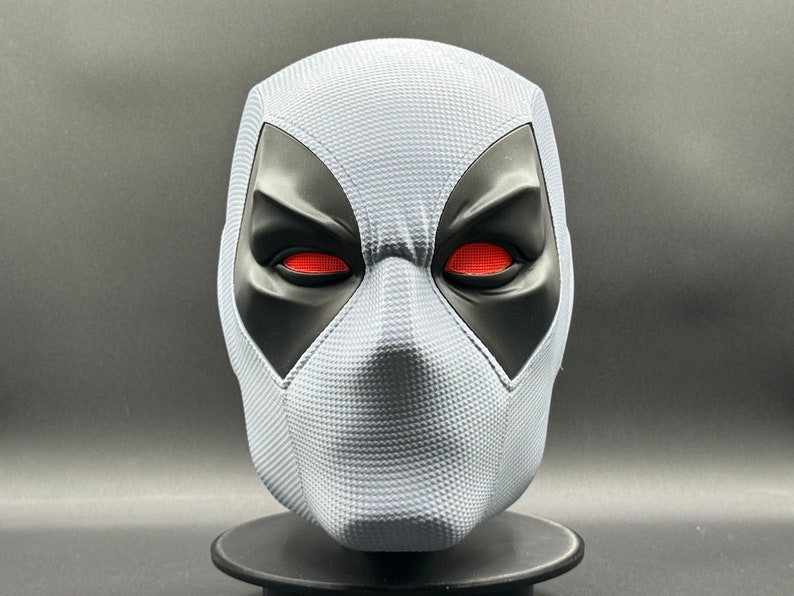 Może przedstawiać: Biała maska Deadpoola z czarnymi otworami na oczy i czerwonymi oczami. Maska ma fakturowaną powierzchnię.