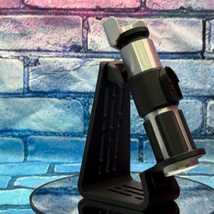 May include: A black display stand holding a silver and black lightsaber. The lightsaber has a silver hilt and black accents. The stand is black and has a unique, angular design. The background is a brick wall with blue, pink, and purple hues.