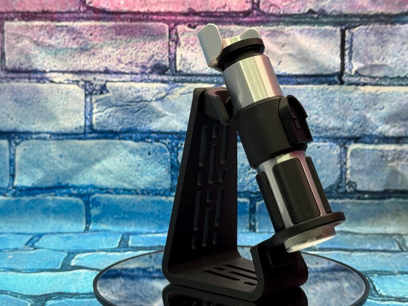May include: A black display stand holding a silver and black lightsaber. The lightsaber has a silver hilt and black accents. The stand is black and has a unique, angular design. The background is a brick wall with blue, pink, and purple hues.