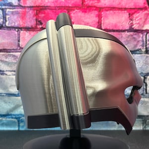 May include: A silver-colored helmet with a dark visor and two cylindrical attachments on the side. The helmet has a sleek, futuristic design, with a dark band around the top and a black base.