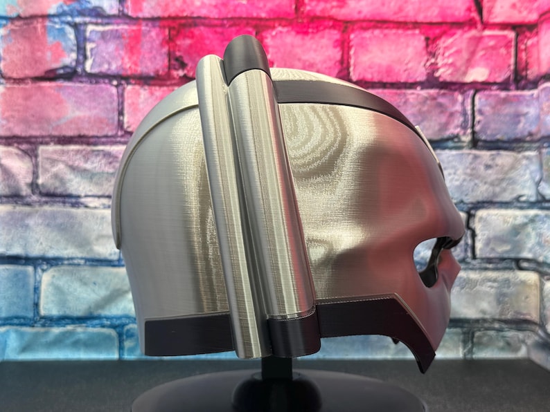 May include: A silver-colored helmet with a dark visor and two cylindrical attachments on the side. The helmet has a sleek, futuristic design, with a dark band around the top and a black base.