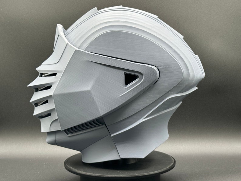 Puede incluir: Un casco impreso en 3D con un dise&ntilde;o detallado, que incluye una visera y una superficie texturizada. El casco es gris y parece estar hecho de pl&aacute;stico.
