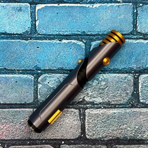 May include: A black and gold lightsaber prop with a cylindrical design. The hilt is dark gray with gold accents, including a ribbed section near the top and circular buttons. The background is a blue brick wall.