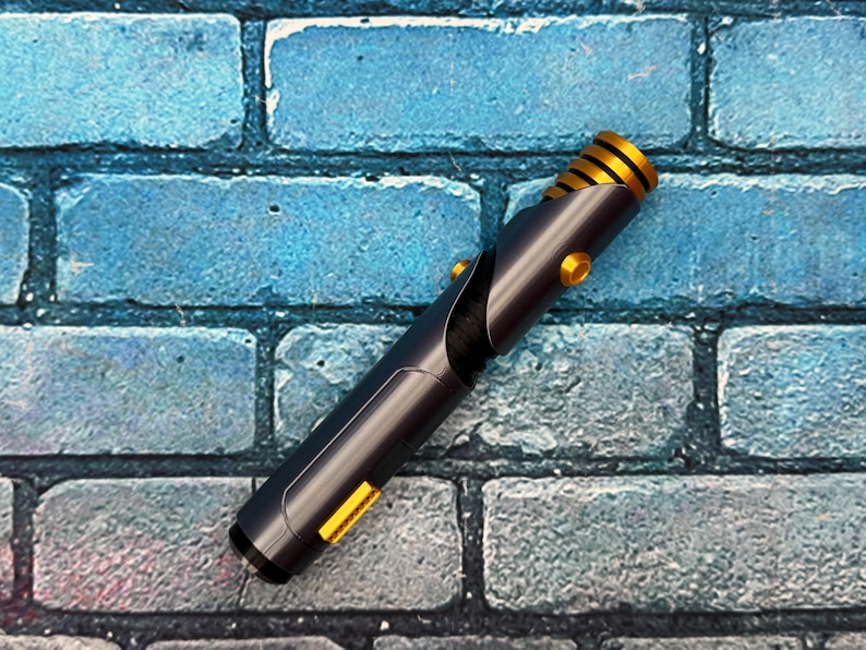 May include: A black and gold lightsaber prop with a cylindrical design. The hilt is dark gray with gold accents, including a ribbed section near the top and circular buttons. The background is a blue brick wall.