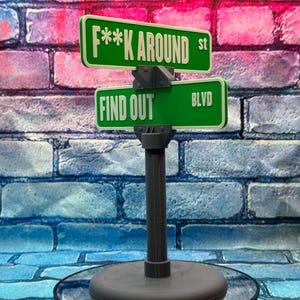 May include: A novelty street sign with green signs that read "F**K AROUND st" and "FIND OUT BLVD" in white lettering. The sign is mounted on a gray post and base, set against a brick wall.