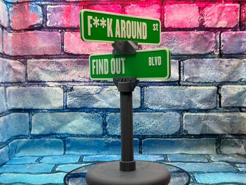 May include: A novelty street sign with green signs that read "F**K AROUND st" and "FIND OUT BLVD" in white lettering. The sign is mounted on a gray post and base, set against a brick wall.
