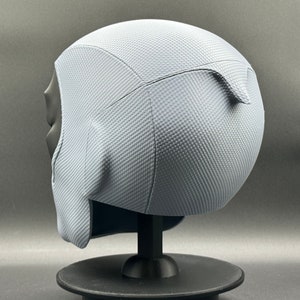 Może przedstawiać: Szary hełm z fakturowaną powierzchnią. Hełm stoi na czarnym stojaku.