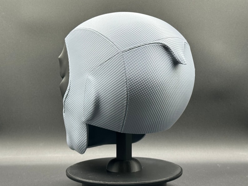 Może przedstawiać: Szary hełm z fakturowaną powierzchnią. Hełm stoi na czarnym stojaku.