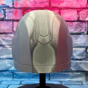 May include: A silver-colored helmet with a smooth, reflective surface. The helmet has a rounded shape with a central ridge and a decorative design on the top. It is displayed on a black stand against a brick wall background.