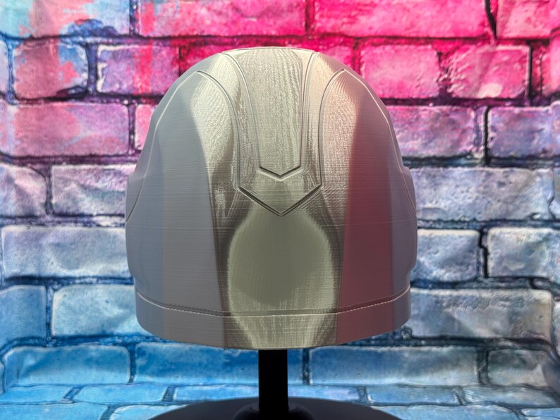 May include: A silver-colored helmet with a smooth, reflective surface. The helmet has a rounded shape with a central ridge and a decorative design on the top. It is displayed on a black stand against a brick wall background.