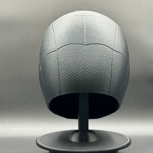 Puede incluir: Casco de pl&aacute;stico negro con una superficie texturizada y un patr&oacute;n de telara&ntilde;a. El casco est&aacute; en un soporte negro.