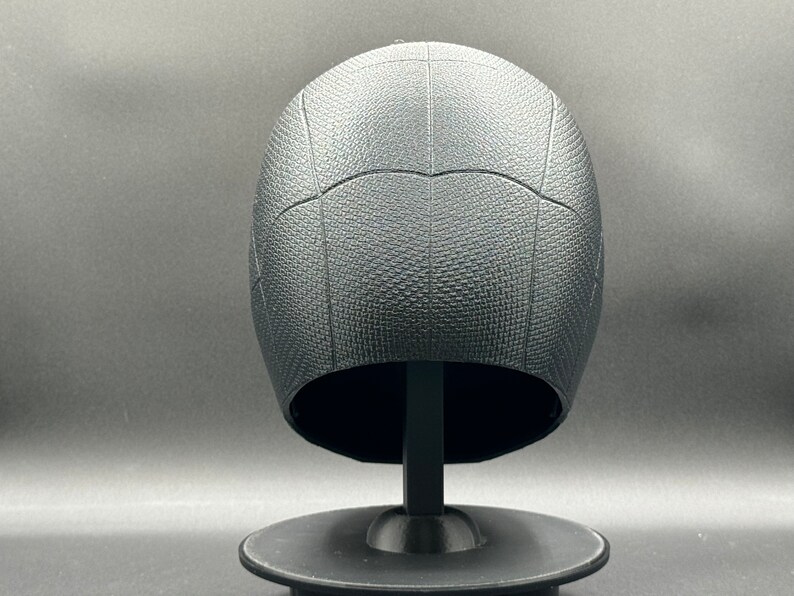 Puede incluir: Casco de pl&aacute;stico negro con una superficie texturizada y un patr&oacute;n de telara&ntilde;a. El casco est&aacute; en un soporte negro.