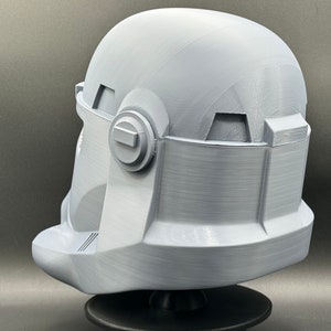May include: A white plastic 3D printed helmet with a textured surface. The helmet has a visor and a round, raised detail on the side.