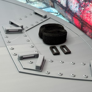 May include: A gray, angular structure with a series of gray rectangular handles and small rivets. Two black hook-and-loop straps and two black rectangular buckles are also present. The background features a colorful, graffiti-style mural.
