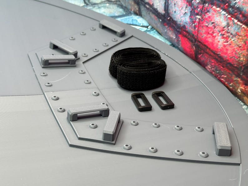 May include: A gray, angular structure with a series of gray rectangular handles and small rivets. Two black hook-and-loop straps and two black rectangular buckles are also present. The background features a colorful, graffiti-style mural.