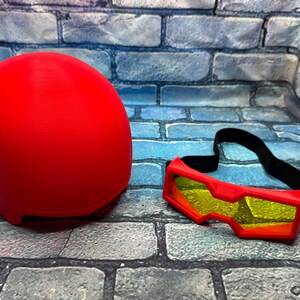 May include: A solid red helmet with a smooth, rounded dome shape paired with a set of red goggles featuring yellow-tinted lenses and an adjustable black elastic strap. The items are presented together as a matching set for head protection.