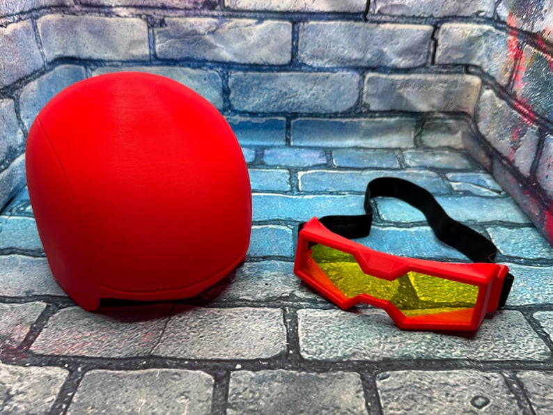 May include: A solid red helmet with a smooth, rounded dome shape paired with a set of red goggles featuring yellow-tinted lenses and an adjustable black elastic strap. The items are presented together as a matching set for head protection.