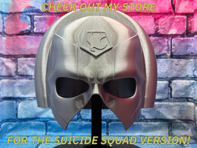 May include: A silver-colored helmet with a distinctive design, featuring large eye openings and a central emblem. The helmet is displayed on a black stand against a brick wall background. Text at the top reads "CHECK OUT MY STORE" and at the bottom "FOR THE SUICIDE SQUAD VERSION!"