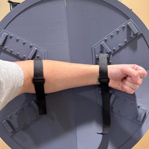 May include: A gray, circular shield with black straps for arm placement. The shield is divided into sections and has metal-like accents. The arm is inserted into the shield, demonstrating its use. The shield appears to be a prop or costume piece.