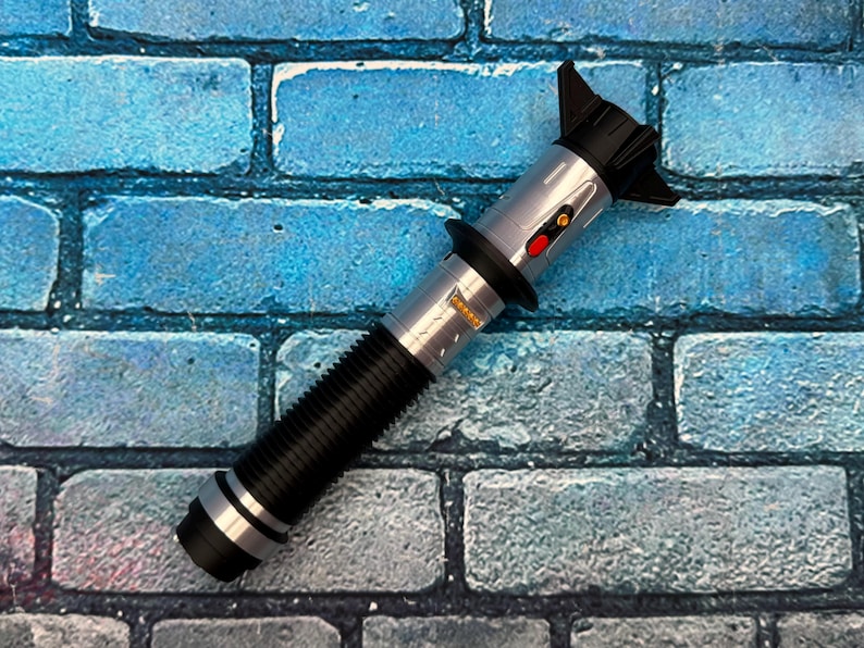 K&ouml;nnte beinhalten: Ein silbernes und schwarzes Lichtschwert-Requisit mit einem geriffelten schwarzen Griff und einem silbernen Emitter. Das Lichtschwert hat einen roten Knopf und ein kleines gelbes Feld. Die Requisite befindet sich vor einer blauen Backsteinmauer.