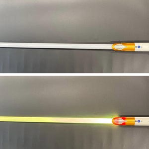 May include: A silver and gold lightsaber with a white blade, and a second image showing the blade illuminated in yellow. The hilt features a detailed design with a silver handle and gold accents. The blade is long and cylindrical.