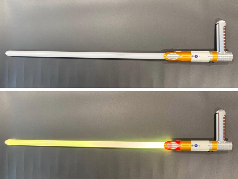 May include: A silver and gold lightsaber with a white blade, and a second image showing the blade illuminated in yellow. The hilt features a detailed design with a silver handle and gold accents. The blade is long and cylindrical.