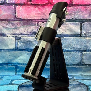 May include: A silver and black lightsaber replica, displayed on a black stand. The lightsaber features a detailed design with silver accents and black grips. The stand is triangular with a slotted design. The background is a brick wall with pink, blue, and purple hues.
