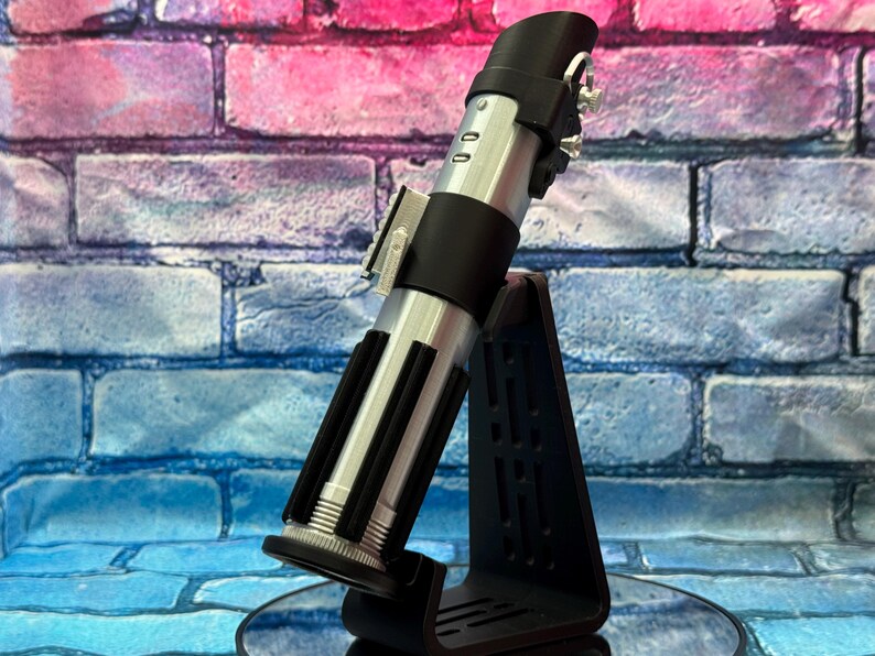 May include: A silver and black lightsaber replica, displayed on a black stand. The lightsaber features a detailed design with silver accents and black grips. The stand is triangular with a slotted design. The background is a brick wall with pink, blue, and purple hues.