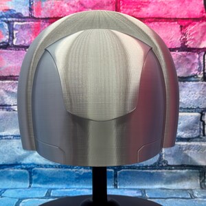 May include: A silver-colored helmet with a rounded top and a visor-like design. The helmet is displayed on a black stand against a brick wall background with pink, blue, and purple hues.