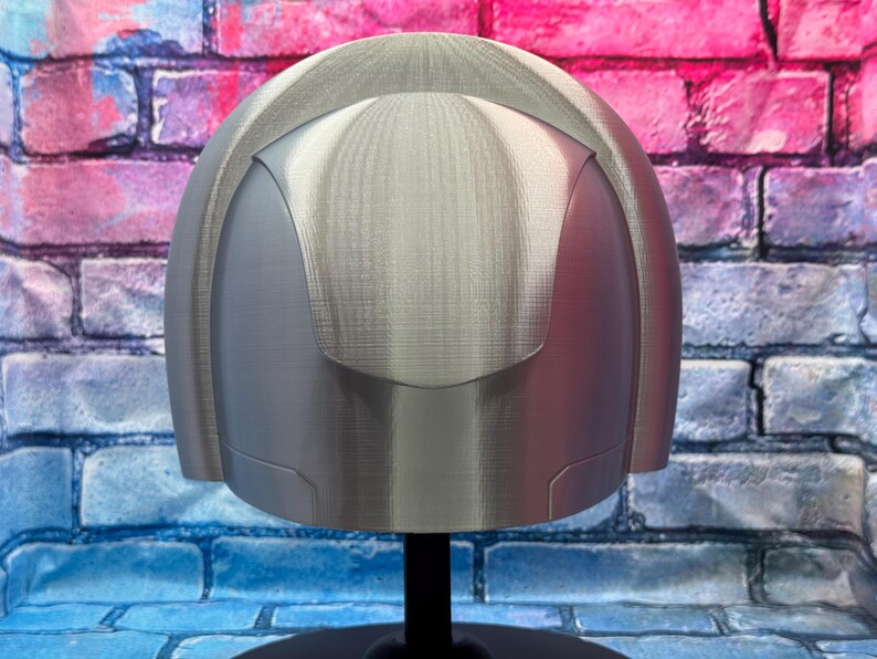 May include: A silver-colored helmet with a rounded top and a visor-like design. The helmet is displayed on a black stand against a brick wall background with pink, blue, and purple hues.