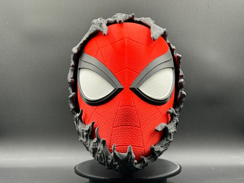 Puede incluir: Una m&aacute;scara de Spider-Man roja y negra con una superficie texturizada. La m&aacute;scara est&aacute; rodeada por un marco negro y dentado.