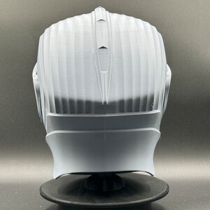 Puede incluir: Un casco blanco impreso en 3D con una superficie texturizada y una parte superior puntiaguda. El casco est&aacute; en un soporte negro.