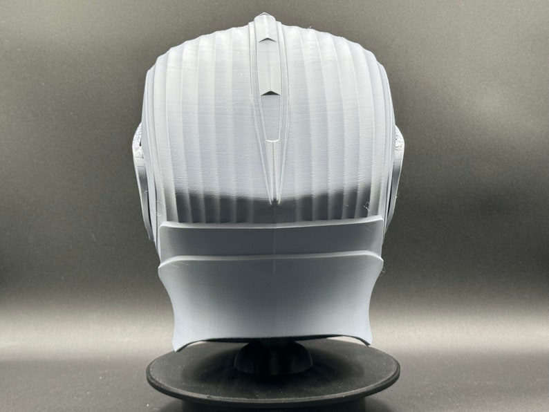 Puede incluir: Un casco blanco impreso en 3D con una superficie texturizada y una parte superior puntiaguda. El casco est&aacute; en un soporte negro.