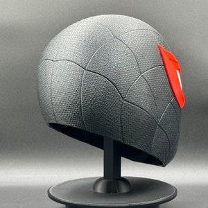 Puede incluir: Casco impreso en 3D negro y rojo con un patr&oacute;n de telara&ntilde;a. El casco est&aacute; en un soporte negro.