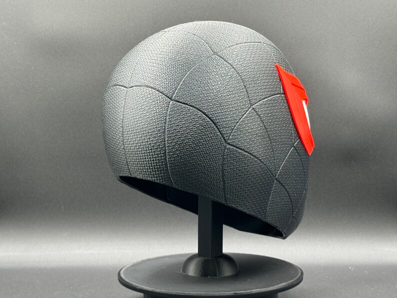 Puede incluir: Casco impreso en 3D negro y rojo con un patr&oacute;n de telara&ntilde;a. El casco est&aacute; en un soporte negro.