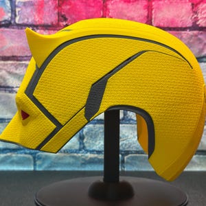 Puede incluir: Un casco amarillo con detalles en negro y gris, con una superficie texturizada. El casco tiene un dise&ntilde;o puntiagudo en forma de oreja y un detalle rojo en forma de ojo. Se exhibe sobre un soporte negro contra una pared de ladrillos.