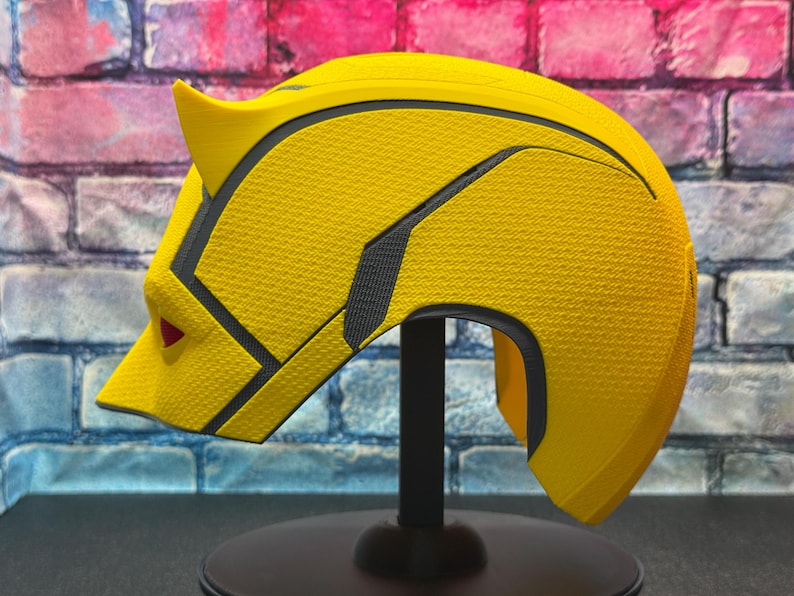 Puede incluir: Un casco amarillo con detalles en negro y gris, con una superficie texturizada. El casco tiene un dise&ntilde;o puntiagudo en forma de oreja y un detalle rojo en forma de ojo. Se exhibe sobre un soporte negro contra una pared de ladrillos.