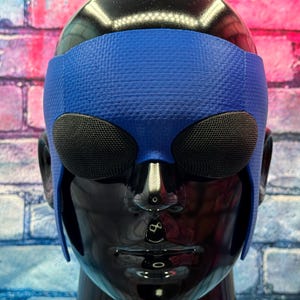 May include: A blue textured headpiece featuring two prominent, oval-shaped black mesh eye coverings. The accessory is fitted onto a glossy black mannequin head, with the blue material extending down the sides to cover the temple and ear areas.
