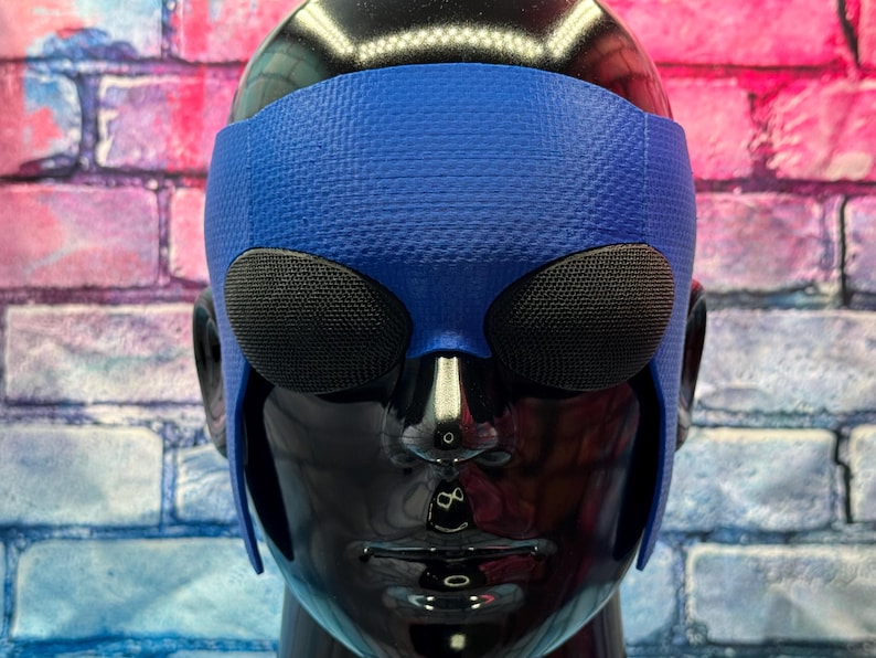 May include: A blue textured headpiece featuring two prominent, oval-shaped black mesh eye coverings. The accessory is fitted onto a glossy black mannequin head, with the blue material extending down the sides to cover the temple and ear areas.