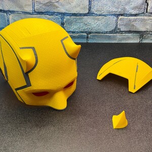 Puede incluir: Un casco de superh&eacute;roe amarillo con aberturas rojas para los ojos y dos peque&ntilde;os cuernos. El casco tiene detalles grises y est&aacute; acompa&ntilde;ado de una visera amarilla separada y una peque&ntilde;a pieza amarilla. Los art&iacute;culos est&aacute;n sobre una superficie oscura.