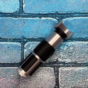 May include: A silver and black lightsaber prop with a detailed design. The hilt is black with silver accents, and the emitter is silver. The prop is set against a blue brick wall background.