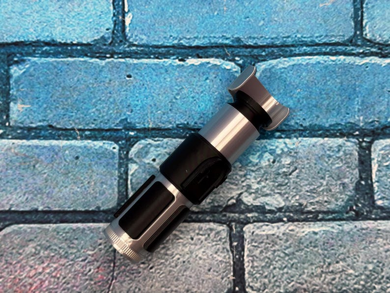 May include: A silver and black lightsaber prop with a detailed design. The hilt is black with silver accents, and the emitter is silver. The prop is set against a blue brick wall background.