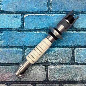 May include: A silver and white lightsaber prop with black accents. The hilt features a white grip with black lines and a silver base. The top has a black emitter and gray details. The prop is set against a blue brick wall.
