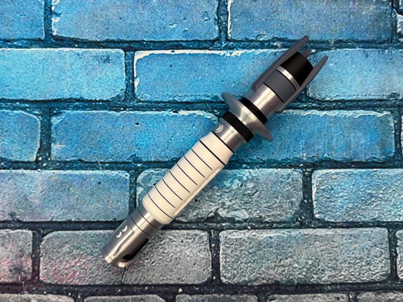 May include: A silver and white lightsaber prop with black accents. The hilt features a white grip with black lines and a silver base. The top has a black emitter and gray details. The prop is set against a blue brick wall.