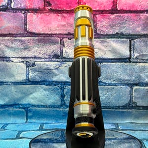 May include: A silver and gold lightsaber prop with black accents, standing upright on a black stand. The lightsaber features a cylindrical design with detailed ridges and a gold emitter. The background is a colorful brick wall.